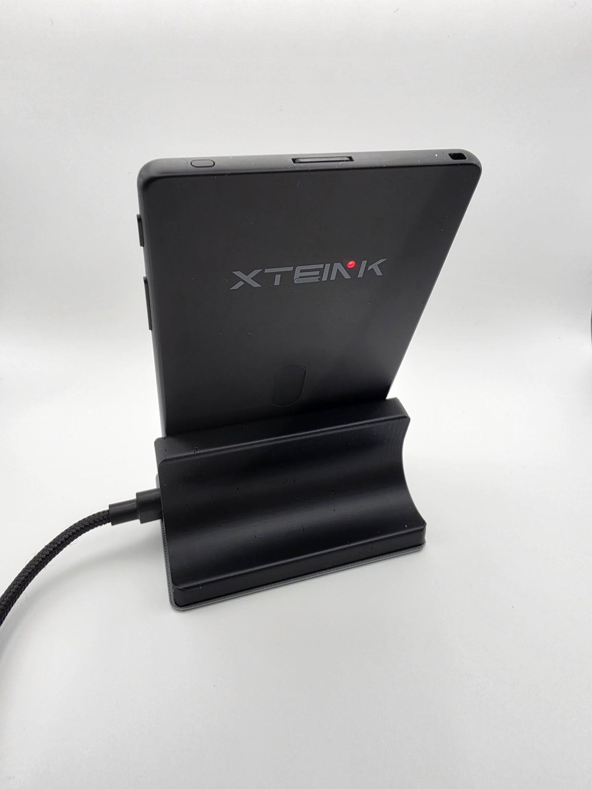 A black XTEINK X4Pro e-bookshown from the back, charging, resting on a matching black charging stand with a cable plugged in, against a plain white studio background.